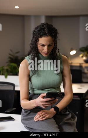 Adult woman sitting on white desk edge, leaning forward, looking at smartphone, wearing green top Stock Photo