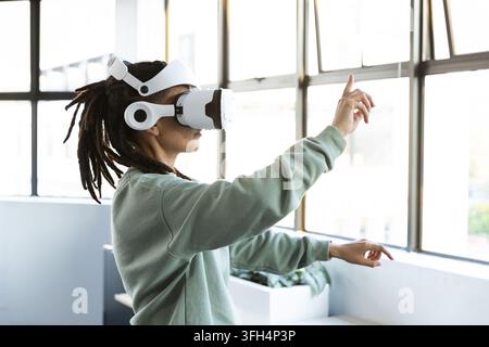 Non-binary person wearing white VR headset and green sweatshirt at studio windows using VR controls Stock Photo