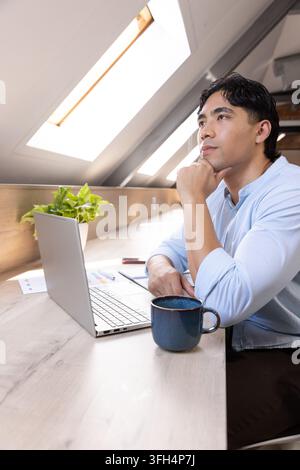 Asian man sitting in loft wearing blue shirt, gazing to skylights, laptop, coffee mug, copy space Stock Photo