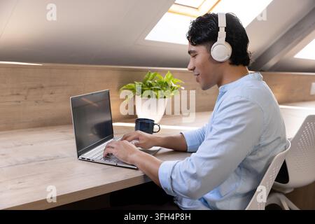 Asian man in 20s working on laptop under skylight wearing blue shirt white headphones, copy space Stock Photo
