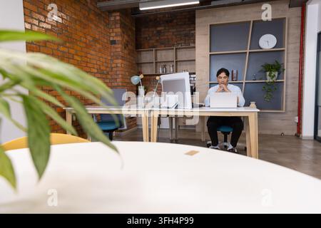 Asian man sitting at desk in office workspace, working on laptop, monitor, wearing light shirt Stock Photo