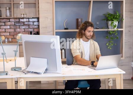 Man in late twenties working on laptop at studio desk wearing beige overshirt, papers, copy space Stock Photo