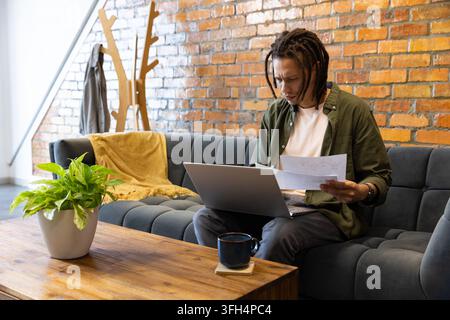 African American man sitting on sofa reading papers while using laptop in lounge, copy space Stock Photo