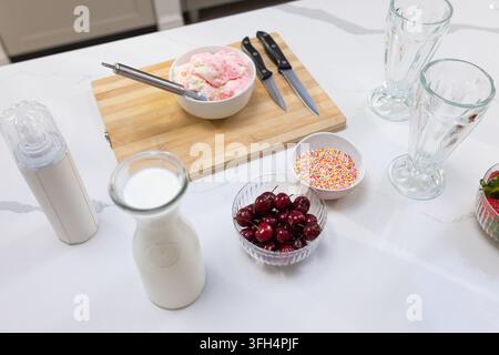 White bowl of pink ice cream is sitting on wooden cutting board, with cherries, sprinkles, glasses Stock Photo