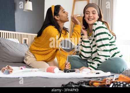 Diverse female friends sitting on bed applying makeup using round mirror and brushes in bedroom Stock Photo