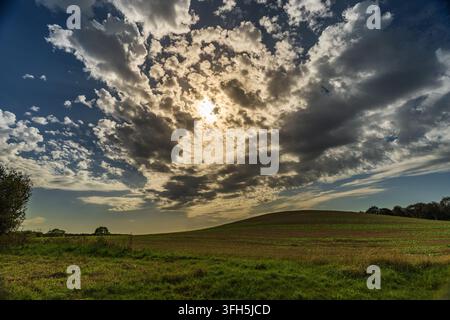 A breathtaking landscape photo capturing the sun breaking through a dramatic, patterned cloud formation, illuminating a vibrant rolling green hill and Stock Photo