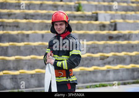 Young firefighter wearing a red helmet and reflective protective gear holds a fire hose on concrete steps, conveying readiness, training, teamwork and Stock Photo