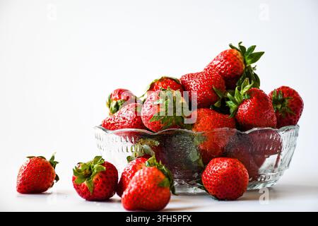 Fresh ripe strawberries piled in a glass bowl with several berries scattered in front, photographed against a bright white background. Stock Photo