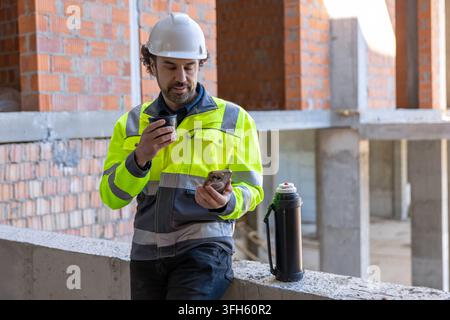 Male architect drinking hot beverage and using smartphone Stock Photo