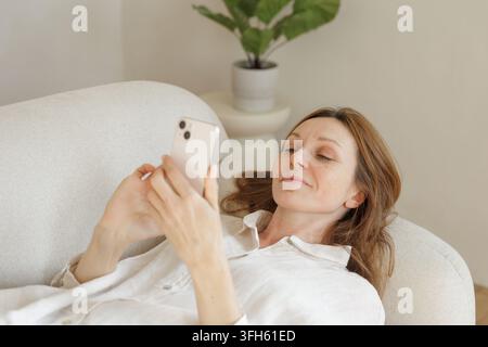 A relaxed woman interacts with an AI voice assistant on her smartphone while reclining on a sofa at home, illustrating conversational technology and e Stock Photo
