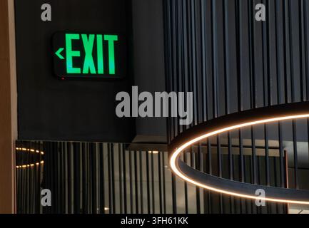An illuminated green "EXIT" sign mounted on a dark wall next to a modern sloped ceiling fixture Stock Photo