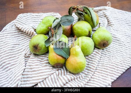Fresh green pears with leaves arranged on striped linen cloth over a wooden table, showcasing ripe seasonal fruit, natural color, and rustic summer ha Stock Photo