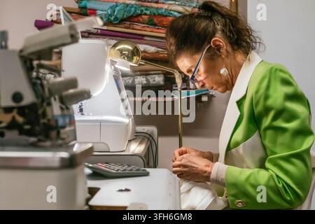 Experienced dressmaker sews a handmade white garment at her workshop table, using a sewing machine, lamp, fabric, thread and tailoring tools for caref Stock Photo