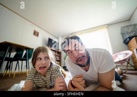Playful father and daughter lying on the living room floor together, pulling silly faces and sharing a humorous family moment at home in natural dayli Stock Photo