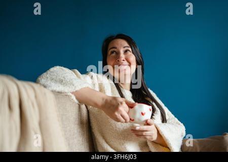 Smiling woman in a cozy white sweater relaxes on a sofa, holding a festive heart-patterned mug against a deep blue wall during the Christmas season. Stock Photo