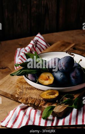 Rustic still life of fresh purple plums with a halved plum, green leaves and striped cloth arranged on a white plate over a wooden table and burlap fa Stock Photo