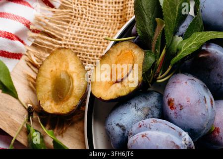 Rustic still life of fresh purple plums on a ceramic plate, featuring two plum halves with visible pits, green leaves, burlap fabric, a wooden board, Stock Photo
