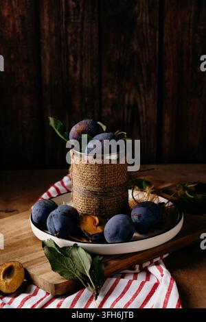 Moody still life of ripe purple plums arranged on a white plate and wooden board, with a rustic metal container, leaves, striped cloth, and dark woode Stock Photo