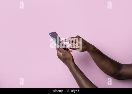 Close-up of black man hands holding and tapping a smartphone against a pastel pink background, representing mobile communication, technology, online s Stock Photo