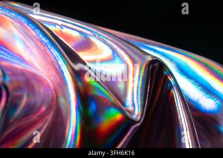 Close-up abstract background of folded iridescent metal with holographic rainbow reflections, glossy curved surfaces, and vivid futuristic light patte Stock Photo