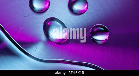 Macro view of clear water droplets resting on an iridescent brushed metal surface, illuminated by vivid cyan and magenta light for a futuristic techno Stock Photo