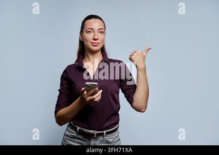 Young businesswoman holding smartphone and pointing thumb to side against grey background Stock Photo