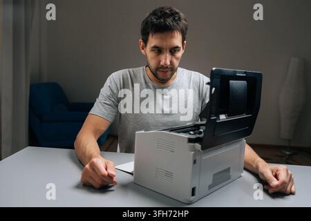 Serious puzzled technician with concentrated expression inspecting laser printer with cover open, attempting to repair it sitting at desk in dark Stock Photo