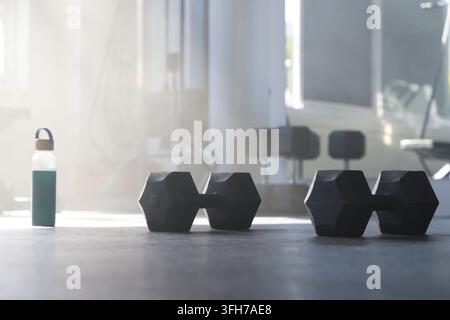 Pair of black hex dumbbells are resting on dark rubber gym floor near green water bottle Stock Photo