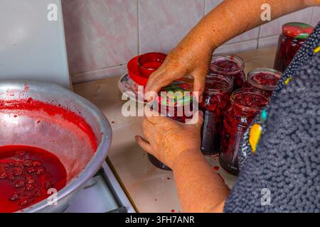 Close-up of hands sealing glass jars filled with homemade strawberry jam. Several jars are arranged on a kitchen counter next to a metal bowl with ber Stock Photo
