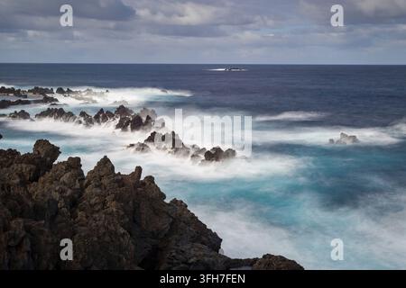 Long exposure captures the roaring sea on a spectacular coast, wild waves break in white spray on rugged, dark volcanic rocks in the lava coast near P Stock Photo