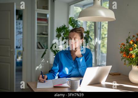 Focused woman working from home sitting at table with laptop, writing plans and ideas in notebook. Stock Photo