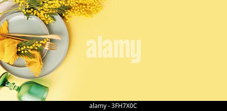 Spring table setting with mimosa flowers and green glass on yellow background with copy space Stock Photo
