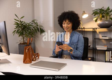 African American woman sitting at office desk holding smartphone with closed laptop, denim shirt Stock Photo