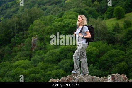 A middle-aged Caucasian woman stands on a rocky outcrop, looking out over a dense, green forest. She is dressed for outdoor activity in a grey t-shirt Stock Photo