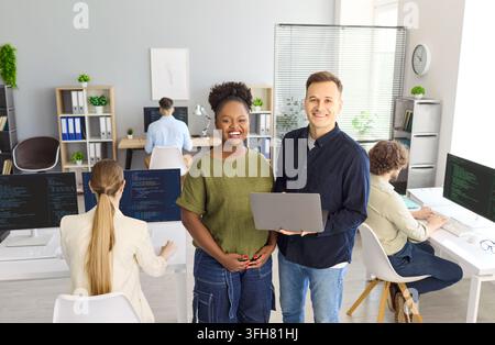 Smiling coworkers with laptop in bright office as programmers code at workstations Stock Photo