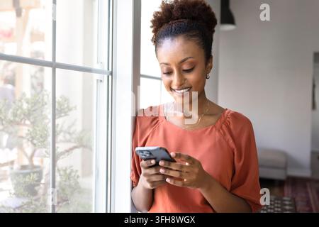 Pretty young woman using modern smartphone standing by window Stock Photo