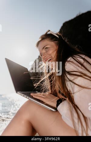 Woman laptop coastal cliffside worker smiles while using her computer at the beach during sunset Stock Photo