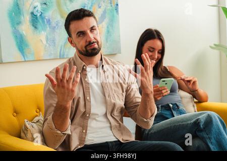 A candid moment captures a man gesturing with open hands while seated on a bright yellow sofa while a woman nearby focuses on a smartphone creating a Stock Photo