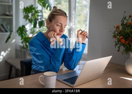 Sleepy pensive businesswoman working on laptop from home sitting at table staring at screen. Stock Photo