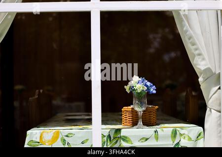 A lemon patterned tablecloth and blue flower vase viewed through bright window with gathered white curtain, creating cheerful outdoor dining setting Stock Photo
