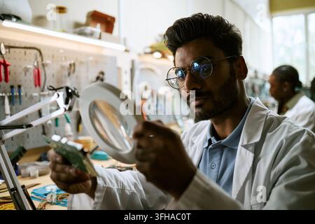 Indian Engineer Inspecting Circuit Board with Magnifier in Tech Lab Stock Photo