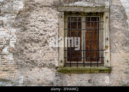 Old window protected by iron bars and framed in stone on a weathered masonry wall, showing aged plaster, exposed brick and traditional architectural d Stock Photo