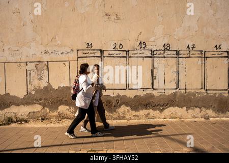 Two people walk along a sidewalk next to a weathered wall with painted window frames. Stock Photo