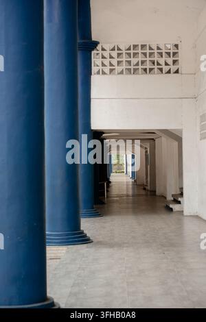 Long empty corridor of a building featuring large blue pillars, white walls, and tiled flooring with vanishing point perspective. Stock Photo