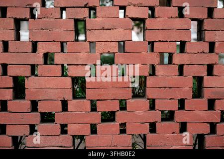Close up of a decorative red brick wall with missing blocks creating a geometric ventilation pattern with green foliage behind. Stock Photo
