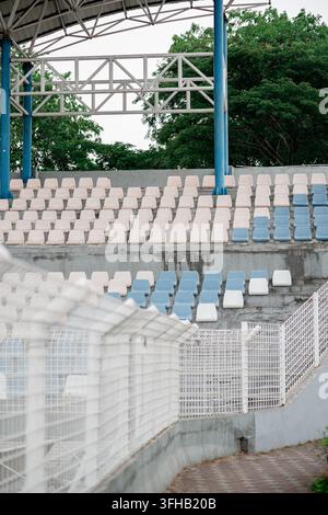 View of empty blue and white plastic seats in stadium grandstand behind a white metal fence. Stock Photo