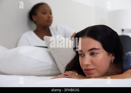 Diverse female friends reclining on bed in softly lit bedroom, background woman holding tablet Stock Photo