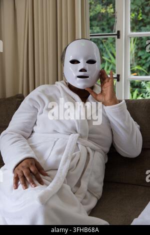 African American female sitting on gray sofa wearing white robe touching LED skincare mask Stock Photo