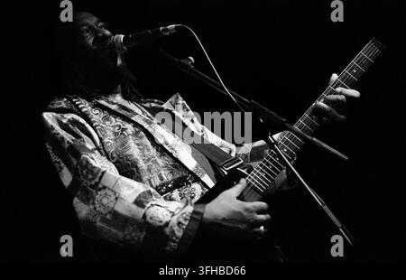 LONDON, UK - FEBRUARY 14, 1994: American avant-garde jazz and free-funk guitarist James Blood Ulmer performing live onstage at Dingwalls nightclub in Camden Lock, London, United Kingdom on February 14, 1994. Ulmer is photographed from a side profile angle, intensely singing into a stage microphone while playing his electric guitar. Dressed in a highly detailed, patterned silk coat, highlighted starkly under deep, high contrast low key concert lighting. This archival monochrome music photograph records a powerful Valentine's Day showcase during his touring era. Stock Photo