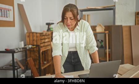 Woman carpenter leaning on table pointing at laptop and reviewing paper plans in workshop; concentration planning. Stock Photo